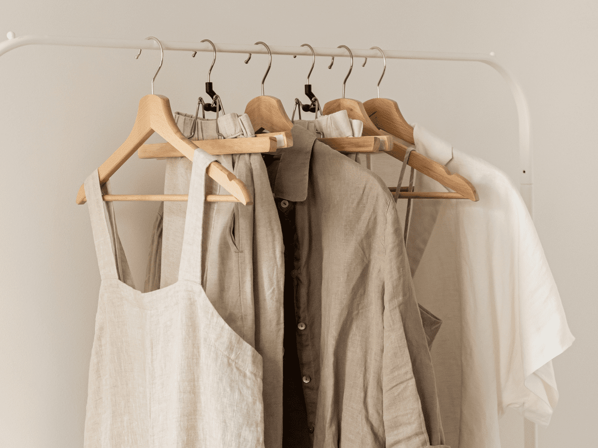 Undyed linen dresses, shirts and an apron in oatmeal, sand and white hang on wooden hangers from a pale rail.