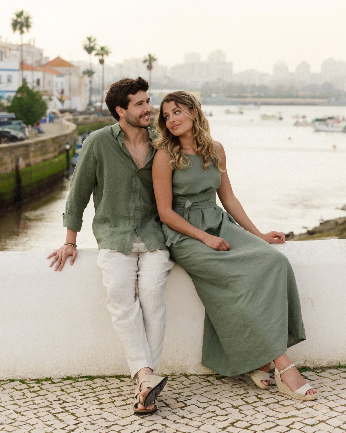 A couple sit on a white harbour wall by the water, she in the sage-green MagicLinen BORACAY long linen dress, he in a sage linen shirt and pale trousers.